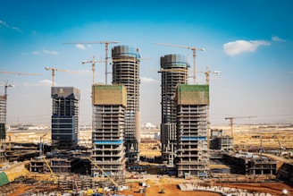 Several high-rise buildings are under construction, surrounded by numerous cranes in a vast, open area. The construction site is busy with scaffolding, partially completed structures, and construction equipment scattered around. The sky is clear with a few clouds, and the scene suggests ongoing urban development.