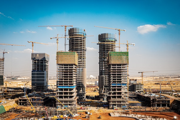 Several high-rise buildings are under construction, surrounded by numerous cranes in a vast, open area. The construction site is busy with scaffolding, partially completed structures, and construction equipment scattered around. The sky is clear with a few clouds, and the scene suggests ongoing urban development.
