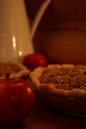 A close-up setting featuring a freshly baked pie with a crumbly topping, accompanied by two red apples. The scene includes a white pitcher in the background, all placed on a wooden surface, creating a warm, rustic atmosphere.