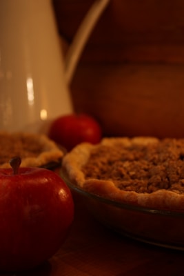 A close-up setting featuring a freshly baked pie with a crumbly topping, accompanied by two red apples. The scene includes a white pitcher in the background, all placed on a wooden surface, creating a warm, rustic atmosphere.