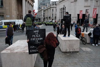 A gathering of people in an urban setting with a visible police presence. A person with red hair is holding a sign with the words 'War is Peace, Freedom is Slavery, Ignorance is Strength.' Several individuals have backpacks, and a bicycle is lying on the ground. Large buildings and flags are part of the background.