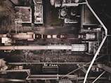 Aerial view of an industrial complex with large pipes and storage tanks. The layout includes a series of interconnected buildings and utilities. The ground and structures appear worn and weathered, indicating prolonged use.