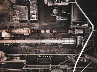 Aerial view of an industrial complex with large pipes and storage tanks. The layout includes a series of interconnected buildings and utilities. The ground and structures appear worn and weathered, indicating prolonged use.