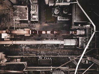 Aerial view of an industrial complex with large pipes and storage tanks. The layout includes a series of interconnected buildings and utilities. The ground and structures appear worn and weathered, indicating prolonged use.