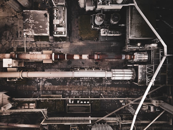 Aerial view of an industrial complex with large pipes and storage tanks. The layout includes a series of interconnected buildings and utilities. The ground and structures appear worn and weathered, indicating prolonged use.