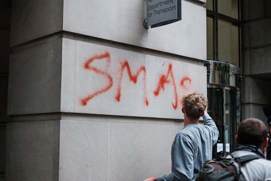 A person with light brown hair and wearing a grey shirt is painting graffiti in red spray paint on the exterior wall of a building. The graffiti reads 'SMASH' on a light-colored stone surface. Another person with short hair and wearing a backpack is nearby. Above the graffiti, there is a sign that indicates this is a governmental department.