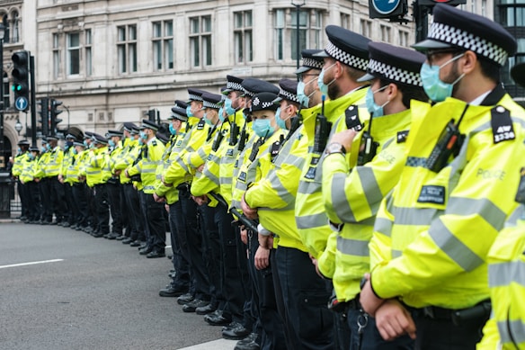 A line of police officers wearing high-visibility yellow jackets and face masks are standing in formation on a city street, positioned near a historic building with large windows. Traffic lights and lamp posts are visible in the background, indicating an urban environment.