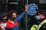 A person wearing a colorful face mask and a red and blue jacket is standing in front of a glass door. They have one hand raised, pressing against a sticker that reads 'Lloyd’s of London' and mentions environmental and social issues. Two police officers in uniform and masks are positioned nearby.