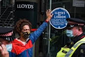 A person wearing a colorful face mask and a red and blue jacket is standing in front of a glass door. They have one hand raised, pressing against a sticker that reads 'Lloyd’s of London' and mentions environmental and social issues. Two police officers in uniform and masks are positioned nearby.