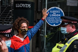 A person wearing a colorful face mask and a red and blue jacket is standing in front of a glass door. They have one hand raised, pressing against a sticker that reads 'Lloyd’s of London' and mentions environmental and social issues. Two police officers in uniform and masks are positioned nearby.