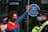 A person wearing a colorful face mask and a red and blue jacket is standing in front of a glass door. They have one hand raised, pressing against a sticker that reads 'Lloyd’s of London' and mentions environmental and social issues. Two police officers in uniform and masks are positioned nearby.
