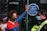 A person wearing a colorful face mask and a red and blue jacket is standing in front of a glass door. They have one hand raised, pressing against a sticker that reads 'Lloyd’s of London' and mentions environmental and social issues. Two police officers in uniform and masks are positioned nearby.