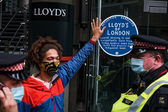 A person wearing a colorful face mask and a red and blue jacket is standing in front of a glass door. They have one hand raised, pressing against a sticker that reads 'Lloyd’s of London' and mentions environmental and social issues. Two police officers in uniform and masks are positioned nearby.