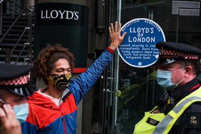 A person wearing a colorful face mask and a red and blue jacket is standing in front of a glass door. They have one hand raised, pressing against a sticker that reads 'Lloyd’s of London' and mentions environmental and social issues. Two police officers in uniform and masks are positioned nearby.
