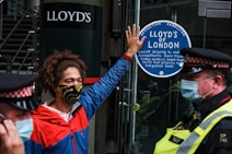 A person wearing a colorful face mask and a red and blue jacket is standing in front of a glass door. They have one hand raised, pressing against a sticker that reads 'Lloyd&rsquo;s of London' and mentions environmental and social issues. Two police officers in uniform and masks are positioned nearby.