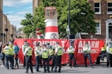 A group of police officers wearing yellow vests stand next to a large red banner that reads 'Sound The Alarm Emergency.' The banner is partially blocking a street and is set up in front of a structure that resembles a large barrel with an 'Extinction Rebellion' symbol. Several people are gathered around, and trees and buildings are visible in the background.