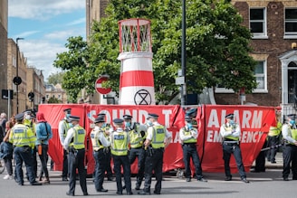 A group of police officers wearing yellow vests stand next to a large red banner that reads 'Sound The Alarm Emergency.' The banner is partially blocking a street and is set up in front of a structure that resembles a large barrel with an 'Extinction Rebellion' symbol. Several people are gathered around, and trees and buildings are visible in the background.