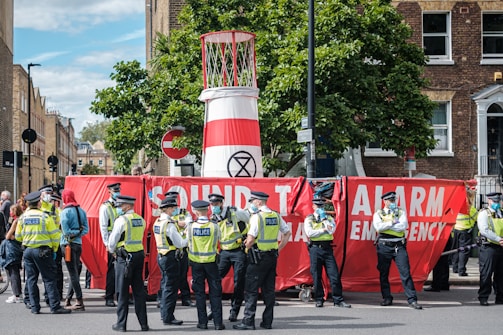 A group of police officers wearing yellow vests stand next to a large red banner that reads 'Sound The Alarm Emergency.' The banner is partially blocking a street and is set up in front of a structure that resembles a large barrel with an 'Extinction Rebellion' symbol. Several people are gathered around, and trees and buildings are visible in the background.