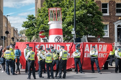 A group of police officers wearing yellow vests stand next to a large red banner that reads 'Sound The Alarm Emergency.' The banner is partially blocking a street and is set up in front of a structure that resembles a large barrel with an 'Extinction Rebellion' symbol. Several people are gathered around, and trees and buildings are visible in the background.