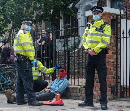 Two police officers in high-visibility jackets and face masks stand over a seated person wearing a red face covering. The scene is outdoors, near a metal fence with a brick wall. Onlookers can be seen in the background, and a bicycle is parked nearby.