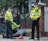 Two police officers in high-visibility jackets and face masks stand over a seated person wearing a red face covering. The scene is outdoors, near a metal fence with a brick wall. Onlookers can be seen in the background, and a bicycle is parked nearby.
