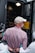A person wearing a white cap and a checkered shirt is handcuffed and being escorted by police officers. The scene appears to be outside, near a police van with an open door, suggesting an arrest is taking place.
