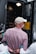 A person wearing a white cap and a checkered shirt is handcuffed and being escorted by police officers. The scene appears to be outside, near a police van with an open door, suggesting an arrest is taking place.
