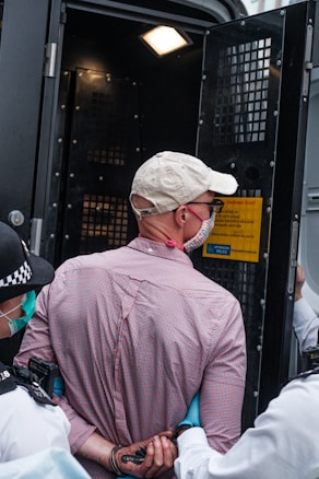 A person wearing a white cap and a checkered shirt is handcuffed and being escorted by police officers. The scene appears to be outside, near a police van with an open door, suggesting an arrest is taking place.