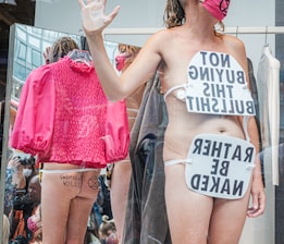 A person stands in a display setting with protest signs that express opposition to consumer culture and fashion. The signs are strategically placed on their body. Reflections in the background show a pink jacket and a group of people, some taking photos. Graffiti on the person's skin reinforces the protest theme.