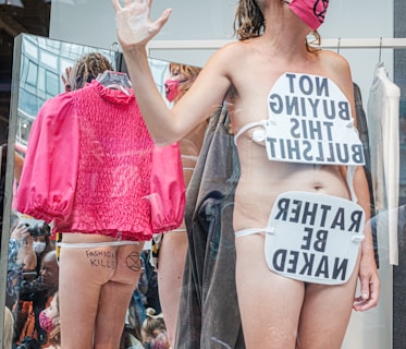 A person stands in a display setting with protest signs that express opposition to consumer culture and fashion. The signs are strategically placed on their body. Reflections in the background show a pink jacket and a group of people, some taking photos. Graffiti on the person's skin reinforces the protest theme.
