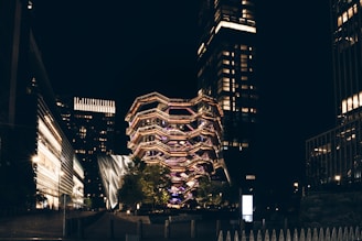 A modern architectural structure illuminated with a variety of lights, surrounded by tall buildings in an urban setting at night. The central structure has a unique, artistic design and is prominently lit.