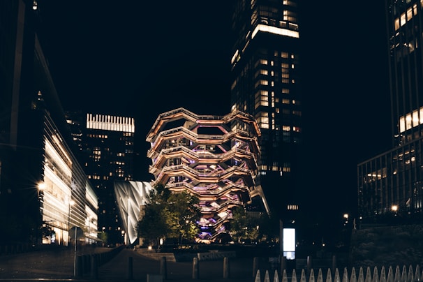 A modern architectural structure illuminated with a variety of lights, surrounded by tall buildings in an urban setting at night. The central structure has a unique, artistic design and is prominently lit.