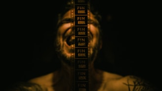 A dark scene where a person is slightly visible in the background with a film strip displaying the word 'FIN' multiple times vertically. The person appears to have tattoos on their shoulder.