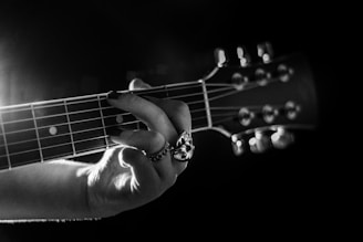 A monochromatic image focusing on the hand of a person playing a guitar. The fingers are positioned on the strings, showcasing a detailed view of the guitar fretboard. The lighting emphasizes the texture of the strings and the intricate design of a ring on the player's finger, creating a dramatic and artistic effect.