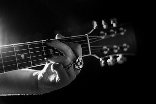A monochromatic image focusing on the hand of a person playing a guitar. The fingers are positioned on the strings, showcasing a detailed view of the guitar fretboard. The lighting emphasizes the texture of the strings and the intricate design of a ring on the player's finger, creating a dramatic and artistic effect.