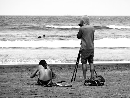 A black and white scene at a beach with a person sitting on the sand, wearing a bikini, facing away, and another person standing with a hoodie, using a video camera on a tripod. Waves roll in the background with a distant person in the water.