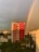A vibrant rainbow arcs over an urban landscape, with a prominent red and white building in the foreground. Surrounding the main building, there is dense green foliage, palm trees, and other residential structures under a dynamic sky with cloud formations.