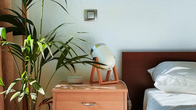 A walnut finish nightstand set beside a bed, holding a warm glowing lamp and a small plant.