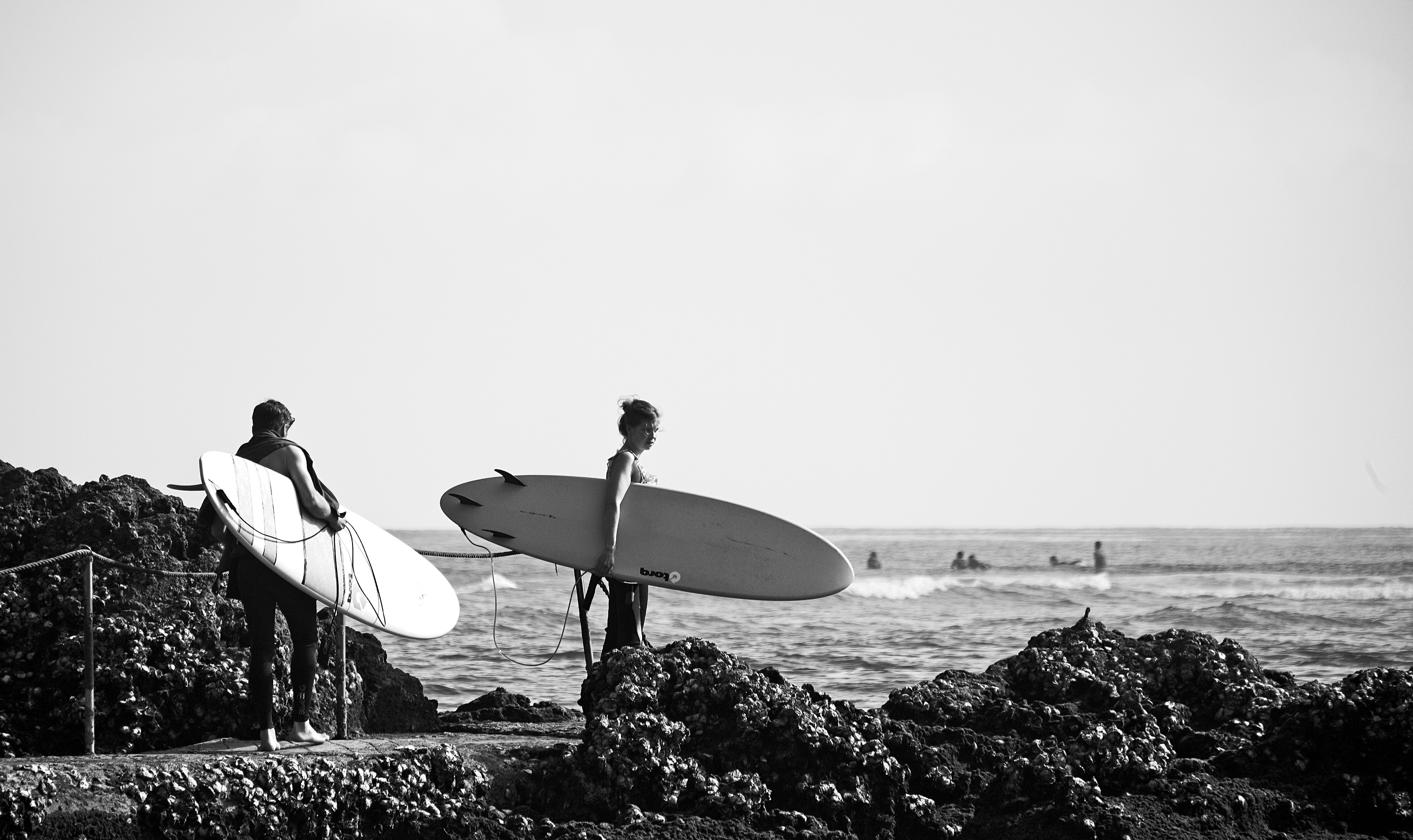 grayscale photo of man holding surfboard standing on rock near body of water