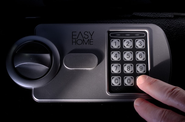 A hand presses the number 0 on an electronic keypad. The keypad is attached to a grey safe with a round handle and the words EASY HOME written above the keypad.