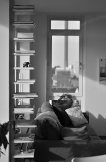 A monochrome interior features a modern shelving unit holding books and a small sculpture of an elephant. A cozy sofa with pillows is placed nearby. Soft natural light filters through a large window that overlooks an urban landscape.