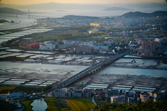 Aerial view of a cityscape featuring a series of rectangular fish ponds or salt pans, surrounded by a mix of residential and industrial buildings. A prominent bridge runs through the middle, connecting different parts of the city across a body of water. The surrounding terrain includes lush greenery, farmland, and distant hills with a river or ocean in the background. The lighting suggests early morning or late afternoon, casting long shadows across the landscape.