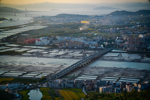 Aerial view of a cityscape featuring a series of rectangular fish ponds or salt pans, surrounded by a mix of residential and industrial buildings. A prominent bridge runs through the middle, connecting different parts of the city across a body of water. The surrounding terrain includes lush greenery, farmland, and distant hills with a river or ocean in the background. The lighting suggests early morning or late afternoon, casting long shadows across the landscape.