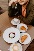 A person is seated at a wooden table with a variety of breakfast items in front of them. The table contains a cappuccino with latte art, two croissants on separate plates, and a dessert featuring a scoop of white ice cream on a bed of berry sauce in a white dish. The person is holding a spoon near their face, dressed in a dark jacket and an orange shirt.