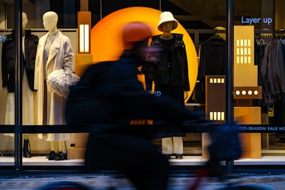 A store window display featuring two mannequins dressed in stylish winter clothing. One mannequin wears a long, white coat and holds a textured handbag, while the other sports a dark outfit with a bucket hat. The background includes geometric designs with bright orange and deep blue colors. A cyclist blurry in motion passes by in the foreground, partially obscuring the view.