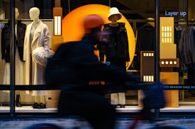 A store window display featuring two mannequins dressed in stylish winter clothing. One mannequin wears a long, white coat and holds a textured handbag, while the other sports a dark outfit with a bucket hat. The background includes geometric designs with bright orange and deep blue colors. A cyclist blurry in motion passes by in the foreground, partially obscuring the view.