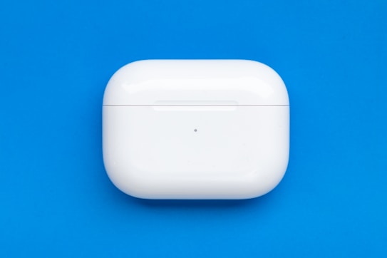 A white, sleek, rectangular case for wireless earbuds is centrally positioned against a bright blue background. The case has a smooth, glossy surface with a small LED indicator on the front.