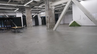 A spacious industrial room with exposed beams and concrete pillars. There's a camping tent set up near the wall on the right, and a few tables and chairs are arranged on the left side. The room has high ceilings with visible lighting fixtures.