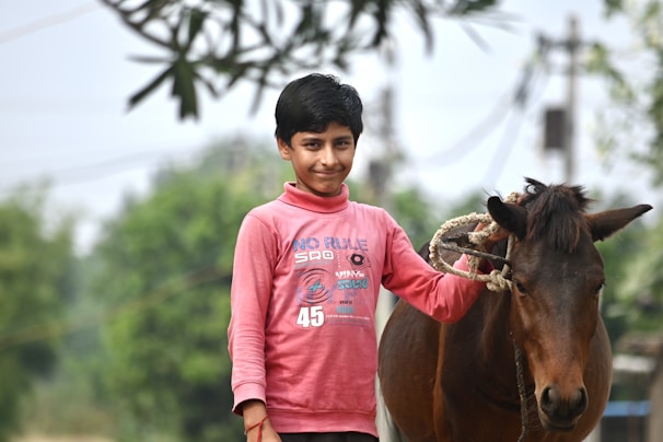 A child holding colorful reins, standing next to a gentle horse under a leafy tree