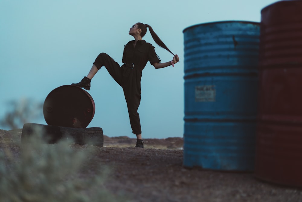 man in black jacket and pants standing beside blue plastic drum